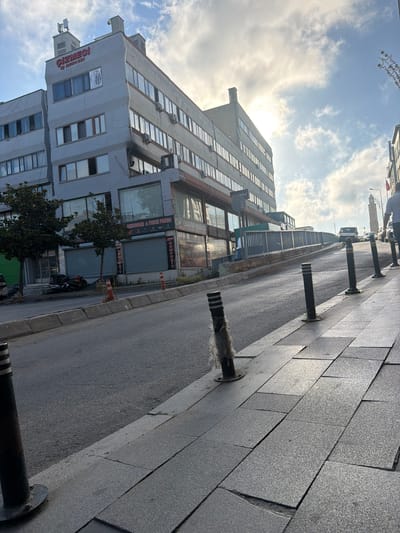 The image shows a street scene in Bayrampaşa, Turkey. In the foreground, a gray paved sidewalk is lined with black bollards. The street slopes upward, with parked cars visible in the distance. On the left side of the street, a multi-story gray building dominates the scene. The building has numerous windows and signage, including "GİZMEGİ İŞ MERKEZİ" in red letters at the top. Trees are planted in front of the building. On the right side of the street, another building with a clock tower is visible further down the road. The sky is partly cloudy with the sun shining through. The overall impression is a commercial street in an urban environment.