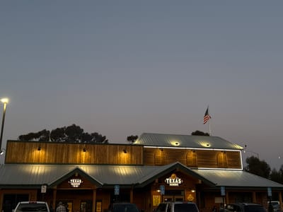The image depicts a Texas Roadhouse restaurant in National City, United States of America, at dusk. The building has a wooden facade with illuminated signs and an American flag waving on top of the roof. Several cars are parked in front of the building. A light post stands to the left of the building. A few people are visible near the entrance. The sky is overcast.
