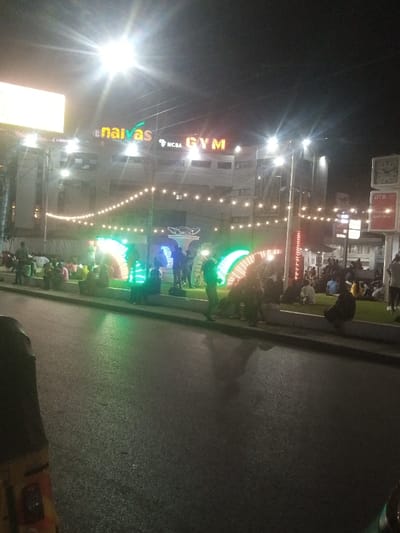 The image presents a nighttime street scene in Majengo ward, Kenya. In the foreground, a dark asphalt road is visible, partially obscured on the left by the front section of a yellow three-wheeled vehicle. Separating the road from a pedestrian area is a concrete median topped with green turf. Numerous individuals are congregated in this pedestrian space; many are seated on the median's edge or directly on the turf, while others stand. The area is brightly lit by string lights draped overhead, prominent streetlights casting significant lens flares, and several large, arched LED light installations emitting green and blue/purple light. In the background, a multi-story building features illuminated commercial signage, including 'naivas' and 'NCBA GYM'. To the right, a pole structure displays a square, dual-faced clock and a sign for 'DTB'.