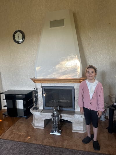 A young girl with blonde hair and a white t-shirt is wearing a pink cardigan and black shorts. She is sitting on a light-colored marble structure in front of a fireplace. The fireplace has a glass door and is surrounded by marble. To the right of the fireplace, there is a black log holder with metal figurines of knights. The room has textured beige wallpaper and brown tiled flooring. A wall clock with a white face and black numbers is visible on the wall. The image appears to be taken indoors, possibly in a living room, during the daytime as indicated by the natural light casting shadows on the wall. The girl has a relaxed expression. There is no visible text in the image.