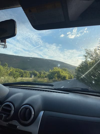 The image, captured from inside a vehicle looking forward through the windshield, depicts a rural road winding through a landscape of rolling hills and lush green trees. The sky is a bright blue with scattered white clouds and what appear to be contrails from an aircraft. Sunlight streams in from the right, creating lens flares and highlighting water droplets or smudges on the windshield. The foreground shows the dark dashboard of the car, including circular air vents. The scene appears to be set in Predgornoe, Kazakhstan, given the context of the prompt.