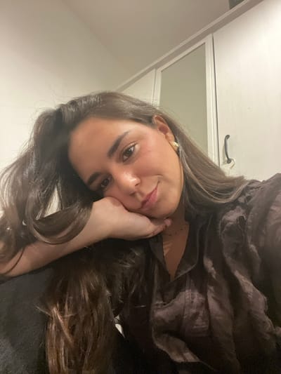 The image displays a head and upper torso shot of a female individual in an indoor setting. She has long, dark brown hair, dark eyes, and is looking directly at the camera with a subtle smile. Her chin rests on her right hand, which is slightly curled. She wears a dark, possibly brown or charcoal, textured collared shirt and a small, golden-colored earring in her visible left ear. A faint dark necklace is also visible. The background consists of a plain, light-colored wall and the upper right portion of a white or light-toned wardrobe or cabinet, which features a mirrored door section and a dark handle. The lighting is soft and appears artificial. The specific location in the city of Bath, United Kingdom, cannot be determined from the visual information provided in the image.