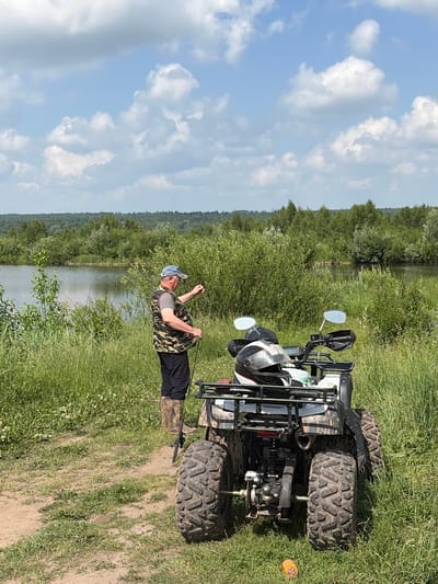 A man wearing a camouflage vest, black pants, and camouflage boots stands near a lake, holding a fishing rod. He is facing away from the camera and towards the water. Beside him is a white all-terrain vehicle (ATV) with a helmet on the back rack. The ATV is parked on a dirt path in a grassy area. The background features a dense line of trees and a cloudy blue sky. The scene appears to be a recreational activity at a lake.