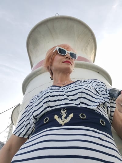 A woman with short, orange hair and white, angular sunglasses stands outdoors, her body angled away from the camera and her head turned to the right, looking upwards. She wears a navy and white striped shirt with a navy band across the chest adorned with a gold anchor and two circular embellishments. Her left arm is visible, resting on a white cylindrical structure that curves over her head, against a bright, overcast sky. A dark cylindrical object is held in her right hand, partially obscured. The woman is positioned in front of a large, white structure with a red stripe, which appears to be part of a lighthouse or a similar beacon. The image is taken from a low angle, emphasizing the scale of the structure and the sky. The setting appears to be Shchetinino, Russia.