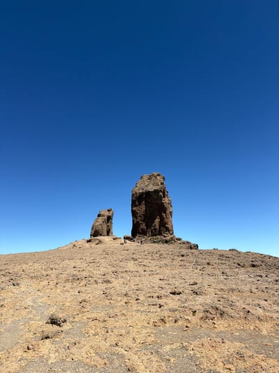 The image shows the Roque Nublo, a prominent rock formation located in Tejeda, Spain. The rock formation is situated on a barren, rocky terrain under a clear blue sky. The foreground is filled with textured ground, composed of small stones and debris. In the mid-ground, the Roque Nublo stands tall, with a smaller rock formation to its left. A few people are visible near the base of the rock formations, appearing small in comparison to the scale of the geological features. The overall environment is arid and rugged, suggesting a mountainous or high-altitude landscape.