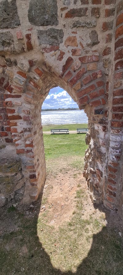The image shows a view through an opening in a brick structure. The brickwork is weathered and set in mortar. The opening reveals a scene with water in the background, followed by a grassy area with two benches. A portion of the grassy area and some soil are visible on the right side of the opening. A shadow is also visible on the right side of the image, falling on the grass. This is likely a photograph taken at the Salaspils Memorial Ensemble in Salaspils, Latvia.