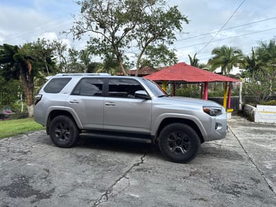 A silver Toyota 4Runner SUV is parked on a cracked, dark grey concrete surface in Quimbaya, Colombia. The vehicle is positioned with its front facing right, presenting its left profile. It is equipped with black multi-spoke wheels, tinted windows, and roof rails. To the left of the vehicle, a sloped green lawn with a low white border transitions into an area of dense tropical foliage and various trees, including one with a white-painted lower trunk. To the right of the vehicle, a red-roofed, open-sided structure with yellow and pink support pillars is visible. Beyond this structure, more tropical trees, including palm trees, and a white, low-lying wall with yellow trim are present. The sky is overcast with visible utility lines stretching across the upper portion of the frame. No individuals are depicted.
