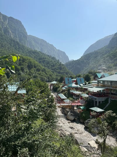 This image captures a scenic view of Qırızdəhnə, Azerbaijan, characterized by a village nestled within a lush, mountainous valley. The foreground is dominated by dense green foliage, with trees partially obscuring a view of a river flowing with muddy, turbulent water. Several large, grey rocks are visible in and around the riverbed.

The village itself features numerous structures with distinctive turquoise roofs. These buildings appear to be restaurants or guest houses, with open-air seating areas, some covered by green awnings. Wooden bridges with red railings span across the river, connecting different parts of the settlement. The architecture varies, with some buildings having a modern, angular design, while others have more traditional A-frame roofs. In the background, towering mountains covered in dense green forest rise steeply, their peaks disappearing into the clear, pale blue sky. The overall atmosphere is one of natural beauty and rustic development. There are no visible people in the image.