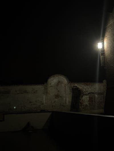 The image depicts an outdoor scene at night in Lagnasco, Italy, likely within the historic grounds of Castello di Lagnasco. A weathered brick wall, partially covered in plaster, forms the backdrop. An arched alcove is visible within the wall, its rough texture suggesting age and disrepair. To the right, a brick building rises, illuminated by a single lamp mounted on its facade. The lamp emits a strong, upward beam of light that cuts through the darkness. In the foreground, a dark, textured surface, possibly a patio or courtyard, is visible, leading to a shadowed area where a single, dark, amorphous shape is situated. The overall atmosphere is one of quiet solitude and historical resonance.