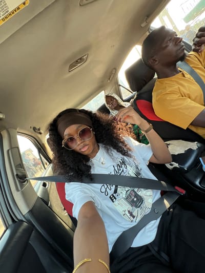 A car interior shows three individuals. In the driver's seat, a person in a white shirt with a graphic and brown headband, wearing rose-tinted glasses, has a seatbelt on. In the back, a person with a white patterned hood looks towards the front, while another person in a yellow shirt is angled away, also wearing a seatbelt. The interior is tan, and a yellow warning sticker is visible on the left. Sunlight streams into the car through the windows.