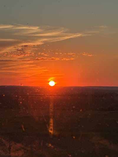 A vibrant sunset paints the sky with hues of orange and red, casting a warm glow over a vast, flat landscape. The sun, a brilliant orb, is low on the horizon, its light diffused by wispy clouds. Below the sky, rolling hills are silhouetted against the fiery backdrop. The foreground is a dark expanse, possibly fields or undeveloped land, dotted with what appear to be blurred lights or reflections. The overall impression is one of natural beauty and serenity, with the setting sun creating a dramatic and colorful display. This scene is observed from a distance, possibly through a window, as indicated by small, out-of-focus circular blurs scattered across the lower portion of the image.