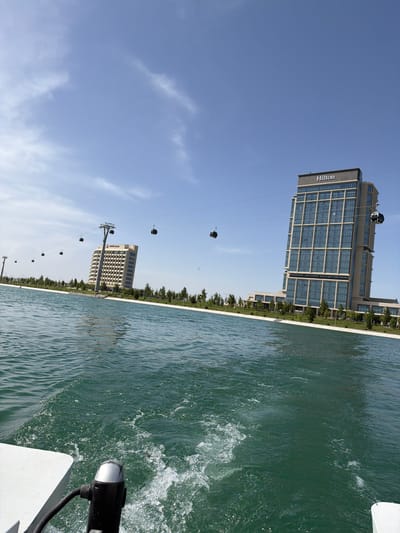This image captures a vibrant waterfront scene in Kuchtumgali, Uzbekistan, viewed from a moving boat. The photograph is oriented sideways, with the horizon appearing vertical.
In the foreground, a body of green-tinted water displays ripples and splashes, indicating movement, likely from the boat the photo was taken from. Part of a white and black object, possibly a boat's hull or motor, is visible in the bottom right corner.
Along the water's edge, a line of green trees separates the water from the land. Beyond the trees, two prominent buildings stand under a clear, bright blue sky with a few wispy clouds. The taller, more prominent building on the left features a modern design with numerous windows, and the word "Hilton" is clearly visible on its facade. Further to the right, another multi-story building, identified by visible text as "Discovery Park," can be seen.
A series of black cable car gondolas are suspended from a cable line that stretches across the middle ground, indicating an aerial transport system or attraction. The bright sunlight suggests it is daytime, likely late morning or early afternoon, with clear weather.