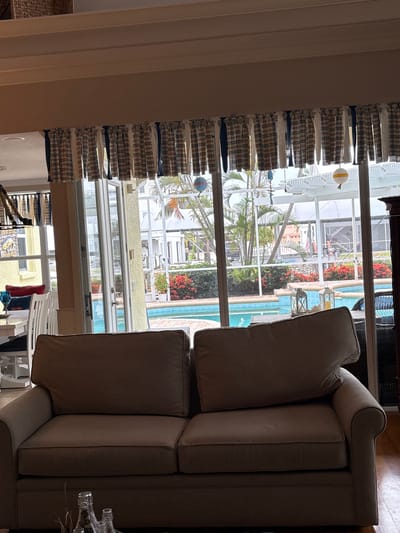 A residential living room features a large, neutral-colored sofa in the foreground, facing a set of glass doors that open to a screened-in patio with a swimming pool. Above the doors is a decorative valance made of alternating plaid and solid navy blue fabric strips. The living room appears to be in Bradenton, Florida, indicated by the typical Florida architecture visible through the glass doors, including palm trees, a pool, and a screened enclosure. The interior decor suggests a coastal or nautical theme, with decorative nautical elements like a hanging buoy and lanterns visible through the glass. The lighting in the room is natural, coming from the outdoor area.