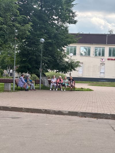 In Kuzmolovskiy, Russia, several people are sitting on park benches in front of a light-colored building. A large tree with green leaves is in the foreground, casting shade. The building has multiple windows and a sign with Cyrillic text. The ground is paved with brick-like patterns. One person is wearing a blue jacket, and others are dressed in casual attire. The sky is partly cloudy.