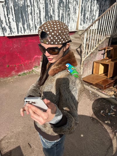 A female individual is depicted outdoors in Neuilly-Plaisance, France, under bright sunlight. She is positioned slightly left of center, her gaze directed downwards at a silver-colored smartphone which she holds horizontally with both hands at waist level. She wears a brown cap adorned with white polka dots, large dark-lensed sunglasses with black frames, a textured jacket resembling brown and grey fur with a distinct brown fur collar, and blue denim jeans. Attached to her right shoulder is a small, brightly colored figurine depicting two parrots: one larger with a blue head and green body, positioned atop a smaller, similarly colored bird.
The background on the left features a red wall exhibiting significant paint degradation and peeling, topped by a section of white wall with heavy peeling and discoloration. To the right, a worn concrete staircase with a corroded white metal handrail ascends. At the foot of these stairs, various wooden debris items, including what appear to be disassembled furniture parts and drawers, are scattered on the ground. The ground is a light grey paved surface, marked by distinct shadows indicating a high sun angle.