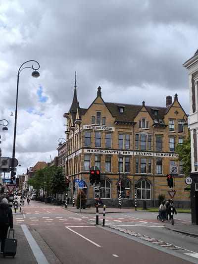 The image captures a street scene in Haarlem, Netherlands, focusing on a yellow brick building with the text "TWEEDE HOLLANDSCHE MAATSCHAPPIJ VAN LEVENS VERZEKERINGEN" visible on its facade. The building exhibits a traditional architectural style with multiple windows and decorative elements. In the foreground, a street with pedestrian crossings and traffic lights is visible. Several people are present, some walking with luggage, others crossing the street. Bicycles are also visible, parked along the street and being ridden in the distance. The sky is overcast with clouds. The overall environment suggests an urban setting with a mix of historical architecture and modern traffic infrastructure.