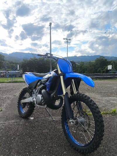 A blue and black Yamaha YZ-series dirt bike (motocross motorcycle) is parked on a dark gray paved surface, leaning slightly to its left on its deployed kickstand. The bike features blue plastic fairings, a black seat, gold-colored front forks, a black exhaust pipe, and knobby tires with blue rims. The paved ground exhibits minor cracking and sparse green vegetation. In the background, from left to right, are distant green, tree-covered mountains, dense foliage, a light-colored building, and blue metallic structures consistent with playground or athletic equipment. Two tall, grey sports lighting poles are visible in the mid-ground. A partial white basketball backboard with a blue rim is present on the far right. The sky is partially overcast with numerous white and grey cumulus clouds. No individuals or active events are depicted. The scene is located in an open area, likely a park or sports complex, within the city of Timaná, Colombia.