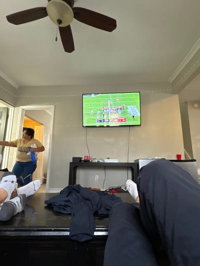 The scene depicts an interior residential space, likely a living room, located in South Bend, United States.
A white ceiling features a brown, four-bladed ceiling fan with a central light fixture. Small, white string lights are affixed along the top perimeter of the light-colored (beige/grey) walls, beneath the crown molding.
On the mid-right wall, a flat-screen television is mounted, displaying an American football game in progress on a green field. The score overlay indicates "35" for the team on the left side of the screen and "40" for "GEORGIA" on the right. Below the television, a black rectangular console table holds a white electronic device, a red Coca-Cola can, and various black cables. Additional cables run from the television to the console table and extend to the floor.
In the foreground, a dark, reflective furniture piece, possibly a coffee table or ottoman, supports the feet and lower legs of at least two individuals. One pair of feet wears grey socks (one marked "CHAMPION") and white socks (one marked "MD"). Another leg, clothed in dark pants, has a white sock. A dark, folded garment rests on this furniture.
To the mid-left, a female individual stands in an open doorway. She has dark hair, wears a light-colored t-shirt with indistinct text, and dark pants. She holds a blue water bottle in her left hand and a white roll of paper or fabric in her right, facing towards an unseen area to her left.
To the right of the console table, a compact silver mini-refrigerator or cooler is present, topped with a red plastic cup. In the far right background, a partial view of a staircase or hallway with light-colored walls is visible.