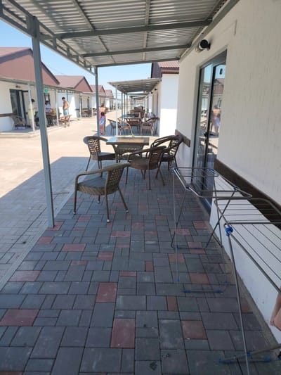 The image depicts an outdoor covered walkway in front of several small, identical bungalows. The walkway is paved with rectangular interlocking bricks in shades of gray and reddish-brown. A metal frame structure with a corrugated metal roof extends over the walkway, supported by vertical metal posts.
In the foreground, several wicker chairs and a table are arranged on the walkway. A metal clothes drying rack stands to the right of the chairs. The bungalows appear to be simple structures with white walls, brown roofs, and large glass doors. Some of these doors have numbers visible on them.
Several people are visible in the background, dressed in casual summer attire, suggesting a vacation or resort setting. Some are standing or walking, while others appear to be sitting at tables outside the bungalows. The sky is clear and blue, indicating a sunny day. The scene suggests a resort or hotel complex in a warm climate. The location is stated to be in Toranghylyk, Kazakhstan.