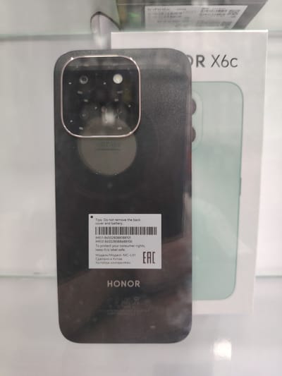 A close-up, slightly angled shot shows the rear of a black Honor X6c smartphone. The camera module is prominently displayed on the top left of the phone's back. To the right of the camera module, the text "OR X6c" is visible on the phone's packaging, which is white. The phone itself is resting on a white surface, possibly a display shelf or table. The background is out of focus, with hints of other electronic packaging or displays. The lighting appears to be bright, likely from overhead store lighting, casting some reflections on the phone's surface. The scene is clean and organized, typical of a retail environment. There are no people visible in the image. The overall impression is a product display. Based on the provided location, this image was taken in Bokhtar, Tajikistan, likely within a mobile phone store or electronics retailer.