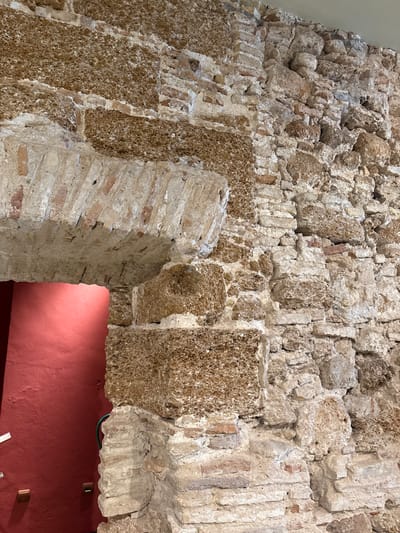 The image depicts a detailed section of an ancient masonry wall and an arched passageway within the Roman Theatre of Cádiz, Spain. The primary wall is constructed from a mixture of irregularly shaped, light-colored, porous stone (consistent with 'piedra ostionera' common to the region) and reddish-brown bricks, all bound by light-colored mortar. The texture is varied, with some sections showing more uniform brickwork and others exhibiting larger, rough-hewn stone blocks.

On the left side, a prominent archway is visible, framed by light-colored bricks set in an angled pattern to form the curve. This arch opens into an interior space characterized by a vibrant, solid red-painted wall. Within this red-painted area, near the lower left of the arch opening, a thin green flexible conduit or wire is partially visible. Further to the left on the red wall, two small, rectangular, light-colored fixtures (possibly electrical outlets or interpretive panels) are mounted vertically. The ceiling above the ancient wall is a plain, light-colored surface. No people are present, and no specific actions or interactions are depicted. The scene highlights the architectural materials and construction techniques of the historical structure.