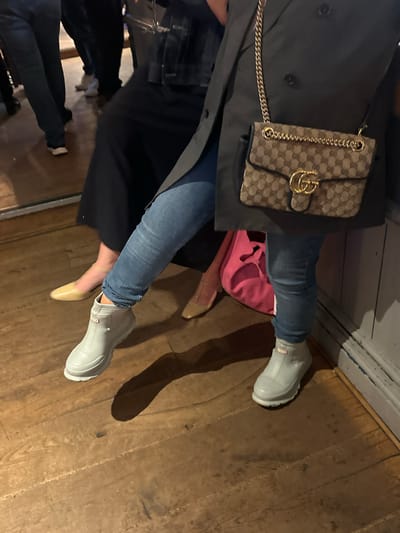 The image depicts two people seated on a wooden floor in what appears to be an indoor setting. One person is wearing blue jeans and light gray Hunter boots, along with a black jacket and a Gucci handbag. The other person is wearing blue jeans and a light-colored shoe, with a black dress or skirt. A pink garment is also visible. The location is St Albans, United Kingdom, based on visual information and other cues. The scene suggests a casual social gathering.