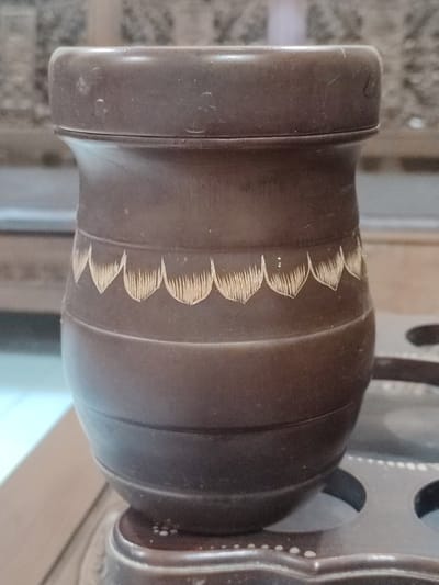 The image shows a close-up, eye-level view of a dark brown, cylindrical object with a rounded lid. The object appears to be made of wood and has a smooth finish. Around the middle of the object, there is a decorative band carved with a repeating pattern that resembles stylized leaves or petals. The carving is light in color, contrasting with the dark wood of the object. The object is sitting on a decorative base, also dark brown, with a patterned edge. The background is blurred, but appears to be an indoor setting with some wooden furniture visible. The lighting suggests it could be daytime, but the overall mood is neutral. No people or text are visible in the image. The location context provided is Blang Cut, Indonesia, which may offer clues about the cultural origin or style of the object.