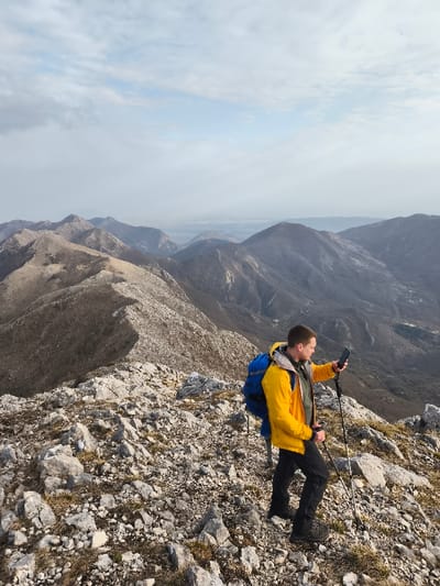 Mali Mikulići, Montenegro: A single male subject, appearing to be in his 20s or 30s, stands on a rugged, rocky mountain ridge. He wears a bright yellow jacket over a darker top, dark trousers, and dark hiking boots, with a royal blue backpack on his back. The subject is holding a black smartphone horizontally in his left hand, looking down at the screen, and gripping a black and silver trekking pole in each hand, with the right pole extended towards the ground. The immediate ground is covered in light grey and white rocks with sparse patches of dry, light brown vegetation. The background features a series of distant mountain ridges and valleys under a hazy, overcast sky composed of diffuse white and light grey clouds.