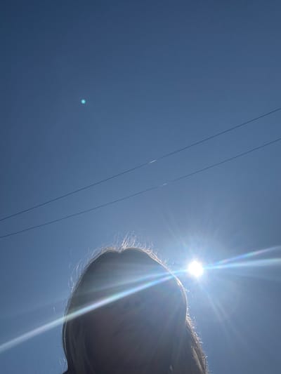 The image is a low-angle shot of a person's head against a clear blue sky. The sun is bright and positioned in the upper right quadrant of the image, causing lens flare that streaks across the frame. Two thin, dark power lines are visible diagonally across the sky. The person's face is obscured by the sun's glare and their hair, which appears light brown and slightly windswept. The overall impression is of a sunny day outdoors, with the sun's intensity creating a dramatic effect. The location is the city of Muravlenko, Russia.
