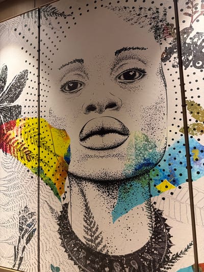 The image shows a multi-paneled artwork, possibly a wall installation, with a central depiction of a human face. The face appears stylized, with a combination of line drawing and stippling techniques. The artwork also incorporates various patterns, including dots, floral motifs, and geometric shapes in black and white, and areas of yellow, blue, and green coloration. The artwork is located in Commune de Diamniadio, Senegal.