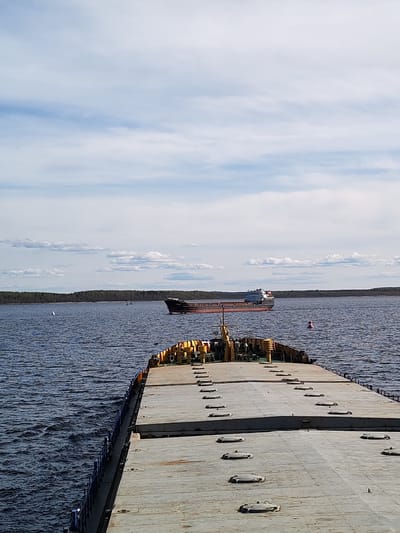 The image depicts a wide-angle shot of a waterway, likely a river or a large lake, in Gorushka, Russia, under a partially cloudy sky. The primary focus is a large, flat-topped barge extending from the foreground into the middle ground. The barge's surface is gray and features multiple access hatches. The railings along the barge are blue.
In the distance, a cargo ship is seen traversing the water. The ship appears to be a general cargo vessel, with a darker hull and lighter-colored superstructure. The background features a low-lying landmass covered in dense forest.
The water is dark blue, reflecting the sky, and there are subtle ripples on the surface. Several navigational markers are visible on the water. The sky above is light blue with scattered white clouds.