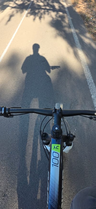 The image shows an overhead view of a bicycle on an asphalt surface, likely a road or bike path. The bicycle's handlebars, front wheel, and a portion of the frame are visible. The frame has "XC 100" printed on it. The shadow of a person holding a camera is cast on the pavement, indicating they are taking the picture. The environment includes a white line on the asphalt and a shadow of trees or other vegetation along the edge of the road. The location is likely a recreational area, potentially a bike path along the coast near the city of Canet d'en Berenguer in Spain.