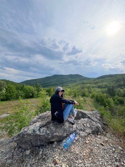 A woman is sitting on a rock outcropping, wearing jeans, a black jacket, sneakers, and a cap. A bottle of water is visible next to her on the rocky ground. Behind her is a green, mountainous landscape under a cloudy sky with the sun visible. The location is near Kamchia, Bulgaria, based on the water bottle label.