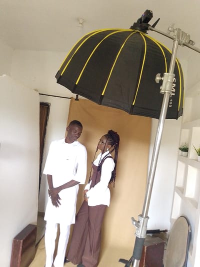 Two young people, a man and a woman, are posing for a photoshoot indoors. The man, on the left, wears a white tunic and trousers. The woman, on the right, is dressed in a white shirt with a brown vest and brown trousers. She has her hair styled in long braids. They are standing in front of a tan backdrop, with a large photography umbrella positioned above them, casting diffused light. A professional lighting stand is visible to the right of the subjects. The background shows a white wall with decorative shelves holding small potted plants. To the left of the subjects, a wooden object leans against the wall. The overall setting appears to be a photography studio. The lighting suggests it is daytime.