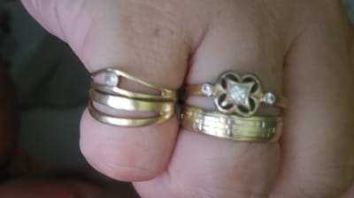 The image shows a close-up of two gold rings on a person's fingers. The ring on the left is a three-banded ring with a small, round, white stone or pearl. The ring on the right is a stack of two rings. The top ring is a decorative ring with a four-leaf clover-like design featuring small diamonds and a small, round, white stone or pearl. The bottom ring of the stack is a thicker, solid gold band with a textured, ribbed pattern. The background is blurred and appears to be a hand or arm. There is no indication of the city of Sasbach, Germany, or any urban environment in the image.