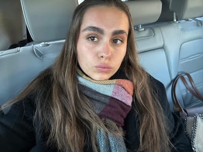 A selfie portrays a woman with long brown hair wearing a dark coat and a multi-colored scarf. She's seated in a vehicle with light gray leather seats. Her gaze is directed towards the camera. Part of a shoulder bag with silver chain detail is visible. The lighting seems bright and natural.