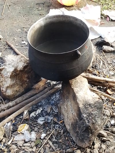 A black metal pot filled with water sits precariously balanced on two rocks over a fire pit. The pot has two handles and a silver rim. The fire pit is made of stones and filled with ash and charred wood. Dry sticks and leaves are scattered around the fire pit. Some plastic waste is also visible on the ground. The scene appears to be outdoors in a rural or semi-urban setting, possibly in Kajola, Nigeria, given the context. There are no people visible in the image.