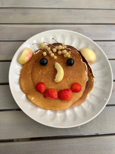 A close-up, top-down shot features a plate of pancakes decorated to resemble a face, set on a wooden outdoor table. The pancakes, stacked two high, form the base of the face. Two slices of peach, positioned on either side of the top pancake, serve as ears. Two blueberries are placed as eyes, above a crescent-shaped slice of banana for the nose. Three strawberries, arranged in a semi-circle below the banana, form the mouth. A sprig of white currants adorns the top of the pancakes. The image is cropped such that the background is not fully visible, but the texture of the wooden table is prominent. There are no people or discernible actions depicted. The setting is likely outdoors, given the lighting and the nature of the table surface. The location is not specified.