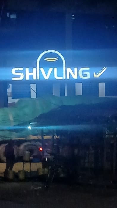 This image depicts a nighttime scene in Kisii Central ward, Kenya. A prominent blue and white illuminated sign displaying "SHIVLING" in block letters, with a stylized arch symbol replacing the 'I' and a checkmark to the right of 'G', dominates the upper portion of the frame. Below "SHIVLING", additional, less illuminated text is partially visible.
In the midground, a light-colored utility vehicle, possibly a pickup truck, is parked, facing right, with its left rear taillight illuminated red. An individual, visible as a dark silhouette, stands near the rear of this vehicle. Several light-colored cylindrical containers, appearing to be buckets, are positioned on the ground and on a low, dark structure next to the individual and the truck. To the right of the individual, a motorcycle is parked upright, mostly obscured by darkness. Behind the parked vehicles and person, a large, dark, mounded mass, possibly covered material or debris, is present. The immediate foreground is a dark, unidentifiable surface, likely pavement or ground. The background is largely dark and indistinct, with a hint of a green tarp or awning behind the lower part of the sign.