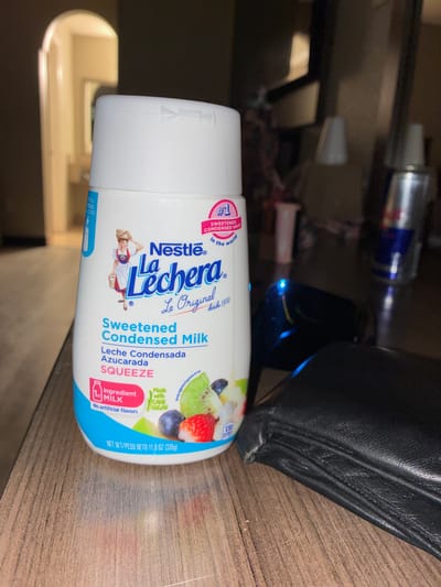 The image shows a close-up of a white plastic bottle of Nestle La Lechera Sweetened Condensed Milk. The bottle is upright and positioned on a wooden surface, likely a table. To the right of the bottle, a black leather wallet is visible, folded and partially obscuring the lower right portion of the bottle. In the background, the scene is dimly lit, with a hint of a doorway or archway on the left, suggesting an indoor environment, possibly a hotel room or a dimly lit restaurant. A red and silver beverage can, possibly Red Bull, is also visible in the background on the right, behind the wallet. The image is taken from a slightly low angle. The lighting appears to be artificial and casts shadows. There are no people or animals present in the image. The overall scene suggests a casual setting, possibly a person’s belongings laid out on a surface. There is no information in the image to suggest the location is in Dallas, United States of America.