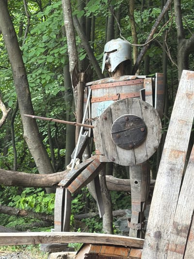 A weathered, life-sized humanoid figure constructed primarily from wood and metal stands upright amidst a dense forest. The figure, resembling a warrior, features a silver-grey, metal helmet with a T-shaped opening for the eyes and mouth, positioned atop a wooden neck. Its torso and limbs are fashioned from varying sizes of wooden planks and staves, secured by dark metal bands and rivets, suggesting a segmented, armored appearance. The figure's right arm (viewer's left) extends forward, holding a thin, unrefined wooden spear. Its left arm (viewer's right) holds a circular wooden shield, composed of multiple planks with a dark, smaller circular boss at its center, secured by additional metal fasteners.
The background consists of numerous mature tree trunks of varying thickness, interspersed with vibrant green deciduous foliage. The ground beneath the figure is partially visible, showing some scattered debris and hints of soil. In the immediate foreground and to the lower right, rough-hewn wooden planks are visible, forming part of an unseen structure or groundwork. The entire scene is set in Golyamo Belovo, Bulgaria.
