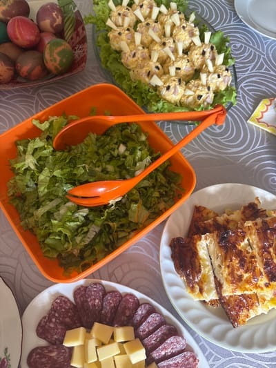 The image shows a table laden with food. There is a salad in a glass bowl with yellow and white toppings and green lettuce. Next to it is an orange bowl filled with green lettuce and a pair of orange serving tongs. Above the salad is a white plate with baked pastries. To the right of the orange bowl is another white plate with sliced salami and cheese cubes. At the bottom left corner is a bowl of painted eggs. The food is arranged on a patterned tablecloth. There is no indication of this being in Klimentovo, Bulgaria. No people are visible in the frame.