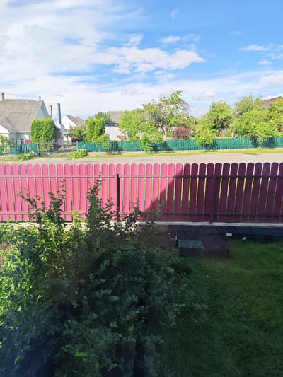 The image depicts a rural residential scene in Ashmyany, Belarus. In the foreground, a dense green bush obscures the lower part of a vibrant crimson metal fence. Beyond the fence, a paved road runs horizontally, with a green picket fence lining the opposite side. Behind the green fence, several houses with pitched roofs are visible, interspersed with lush green trees and hedges. The sky above is a bright blue, dotted with fluffy white clouds, suggesting a pleasant, sunny day.
The scene appears to be a typical residential neighborhood, likely a suburban or village area. There are no people visible in the frame. Scattered on the ground in front of the crimson fence are several black plastic crates and some green plastic sheeting, possibly indicating recent gardening activity or preparations for construction. The time of day is clearly daytime, as evidenced by the bright sunlight casting shadows, particularly on the crimson fence. The overall impression is one of a tranquil, well-maintained outdoor environment under a clear sky.