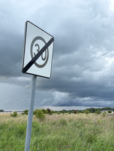 The image depicts an end of speed limit sign with "30" inside a circle, crossed by a diagonal red bar, indicating the end of a 30 km/h speed limit. The sign is mounted on a grey metal pole in a grassy field under a dark, cloudy sky. In the background, a row of white houses with dark roofs is visible, suggesting a suburban or rural residential area. The foreground consists of tall, green grass and some scattered bushes. The overall atmosphere is overcast and slightly ominous due to the heavy clouds. The location is identified as Wilczyce, Poland. There are no people or animals visible in the frame.