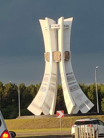 The image captures a monument located in the vicinity of Чарнаўчыцкі сельскі Савет, Belarus. The monument is a tall, white, abstract structure with curved, flared sections at the base and top. It features decorative patterns, possibly of ethnic or traditional design, along its surfaces. Two shield-like emblems are affixed to the central part of the monument.
In the background, a dark, cloudy sky looms over a dense line of green trees. To the left and right of the monument, streetlights are visible. In the foreground, a road is present, with a white car partially visible on the right side, and another vehicle on the far left with its red taillight illuminated. A directional traffic sign with red and white markings is also visible near the base of the monument, indicating a turn. The ground surrounding the monument is covered in a textured, yellowish-brown material, possibly grass or decorative landscaping. The overall scene suggests an outdoor public space, likely a roadside monument or a landmark.