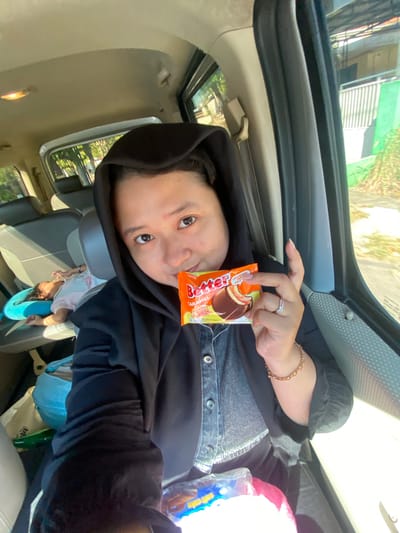 A woman in a black hijab and jacket is taking a selfie in the passenger seat of a vehicle. She is holding an orange and white packaged snack. To her left, a baby is sleeping in a car seat. The car appears to be in motion, with trees and buildings visible through the window. The scene is likely in Randubango, Indonesia, given the context. The woman is wearing a ring on her left hand and a bracelet on her right wrist. The overall lighting suggests it is daytime, with sunlight streaming into the vehicle.