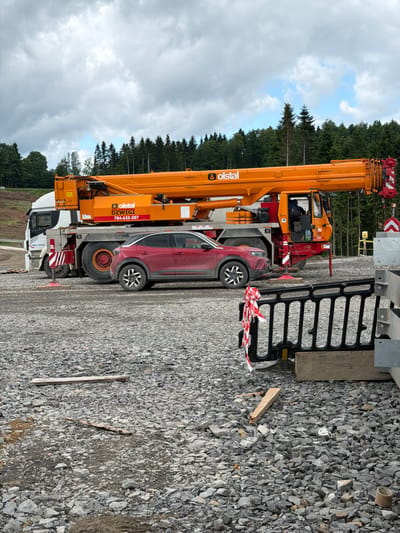 The image shows a construction site in Jodłownik, Poland, on a cloudy day. A large orange crane truck, identified by "olstal" and "DZWIGI" branding, is parked on a gravel surface. A red compact SUV is positioned in front of the truck's rear wheels. The background features a dense forest of evergreen trees and a partly cloudy sky. The foreground is dominated by loose gravel, with some wooden planks and construction barriers visible. A person is seated in the driver's cab of the crane truck.