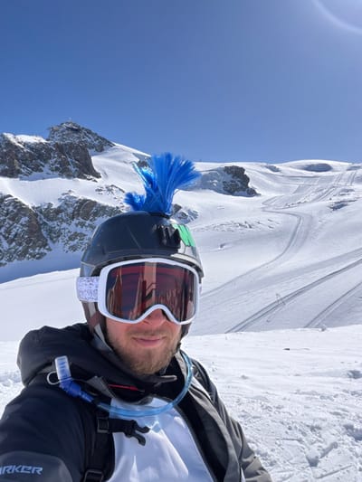 A male individual is depicted in a close-up, chest-up view on a snow-covered mountain within the high-alpine ski domain accessible from Valtournenche, Italy. He wears a black ski helmet featuring a bright blue faux-mohawk attachment and white-framed ski goggles with highly reflective reddish-orange lenses. A green marking is visible on the helmet's side. The individual has facial hair and a slight smile. He is dressed in a dark-colored ski jacket over a white layer, with a blue hydration pack tube and bite valve extending from his right shoulder across his chest. A "MARKER" logo is present on the lower left sleeve of his jacket. The background consists of extensive snow-covered mountain slopes with visible ski tracks. To the left, a prominent rocky peak with a building structure, consistent with a mountain lift station, is visible at its summit. The sky is clear and blue.