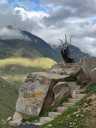 The image depicts a mountainous landscape with a stone sculpture atop a set of stairs. The sculpture is composed of a large, dark, irregular-shaped stone balanced on a stone pedestal, with several metal rods protruding from its top. The stairs are made of stone and concrete, with some overgrown vegetation. A large rock in the foreground features a yellow marking and the numbers "65450" painted in a darker color. In the background, a lush green mountain slopes down into a valley, with a patch of sunlight illuminating a section of it. The sky above is cloudy, with patches of blue visible. The scene appears to be located in Gramphu, India, as indicated by the context provided. There are no people or animals visible in the image.