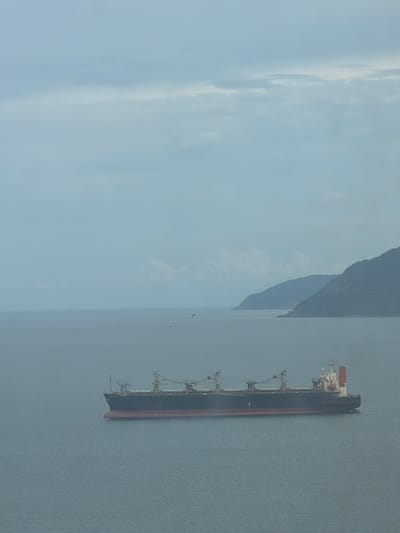 An elevated, wide shot captures a vast expanse of overcast ocean meeting a distant, misty coastline. In the foreground and mid-ground, a large, dark cargo ship with a red waterline is the primary subject. The vessel has several cranes visible on its deck. The sea is calm, with subtle ripples. In the background, two large, dark, mountainous landmasses are visible, their forms softened by the atmospheric haze. The sky is a uniform light gray, indicating cloudy or foggy conditions. No people are visible in the frame. The scene appears to be set in a coastal area, potentially near a port or along a shipping route. Given the description of a cargo ship and mountainous coastline, it is plausible that this image was taken in the vicinity of Hải Vân Ward, Vietnam, which is known for its scenic coastal mountains and proximity to the sea.